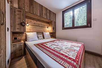 A charming chalet-style bedroom features a bed with a vibrant blanket, rustic wood built-ins, and a window overlooking a forest.