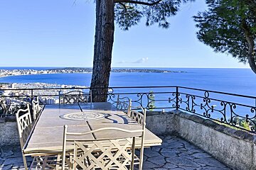 Sunny patio with outdoor dining, a tree, and a wrought-iron railing, overlooking a panoramic blue sea and coastal city.