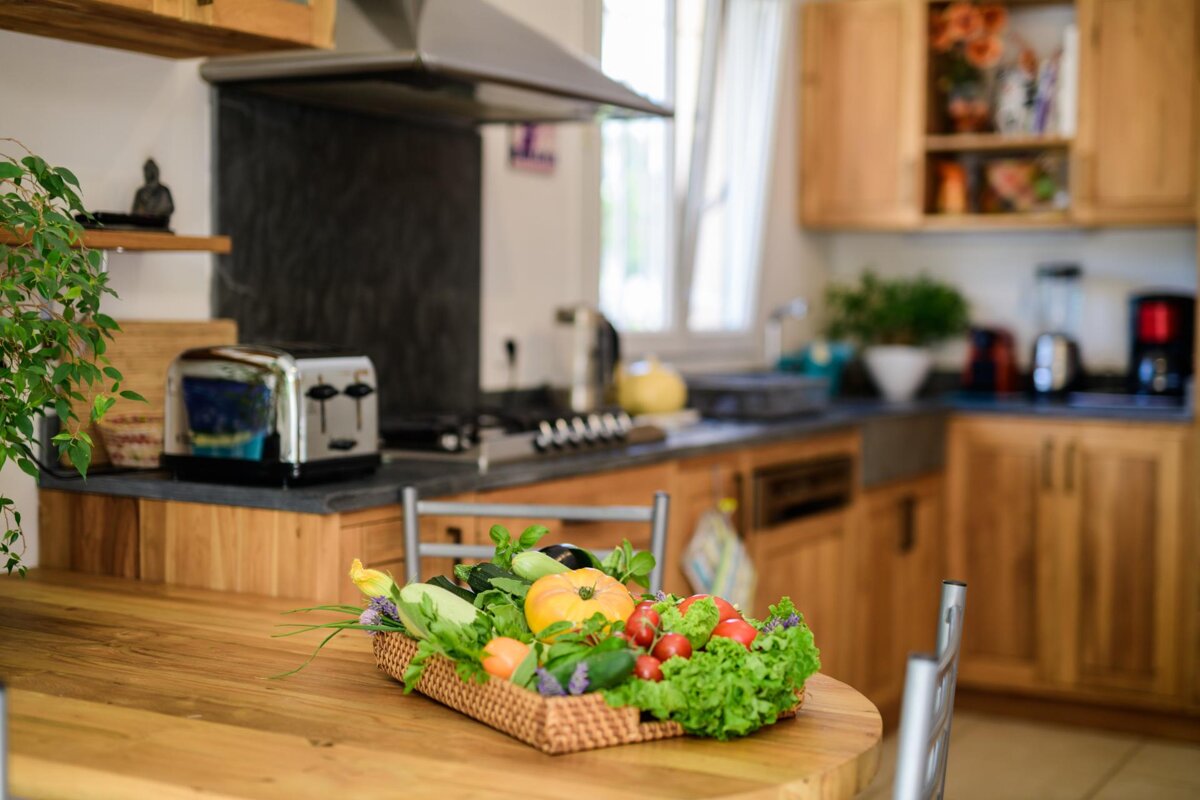 A basket of vegetables sits on a wooden table in a kitchen