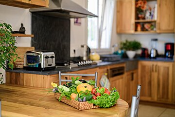 A basket of vegetables sits on a wooden table in a kitchen