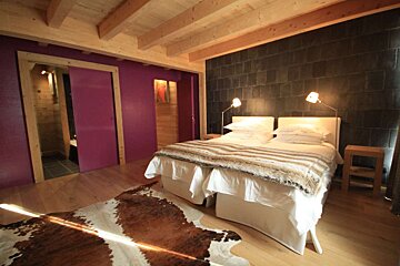 Cozy bedroom with a large bed, cowhide rug, wood floor, exposed beams, dark stone wall, and a bright purple wall with a sliding door.