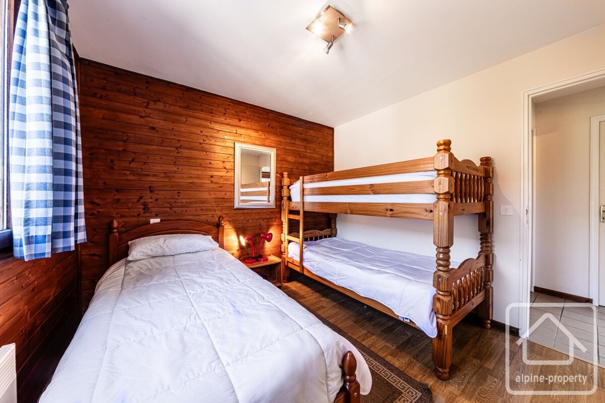 Rustic, wood-paneled bedroom featuring a single bed and a wooden bunk bed with white linens. A blue checkered curtain hangs by the window.