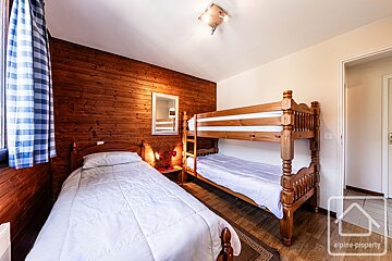 Rustic, wood-paneled bedroom featuring a single bed and a wooden bunk bed with white linens. A blue checkered curtain hangs by the window.