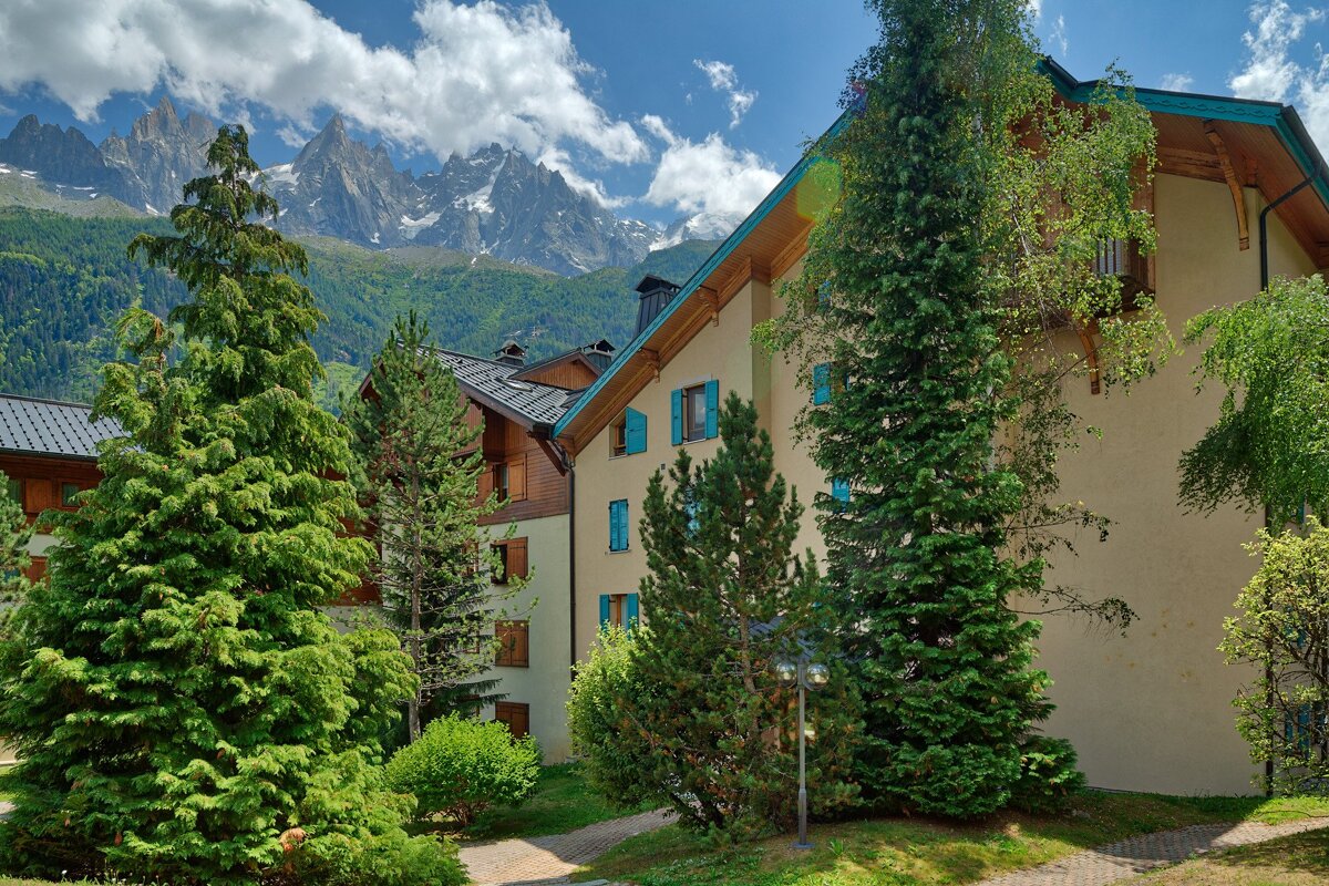 Lodge-style buildings with green trees in front of jagged, snow-capped mountains under a bright, cloudy blue sky.