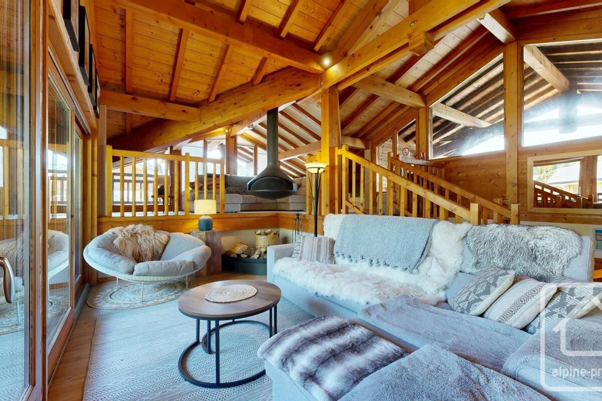 Cozy alpine chalet living room with exposed wooden beams, plush sofas, fuzzy blankets, and a suspended black fireplace.