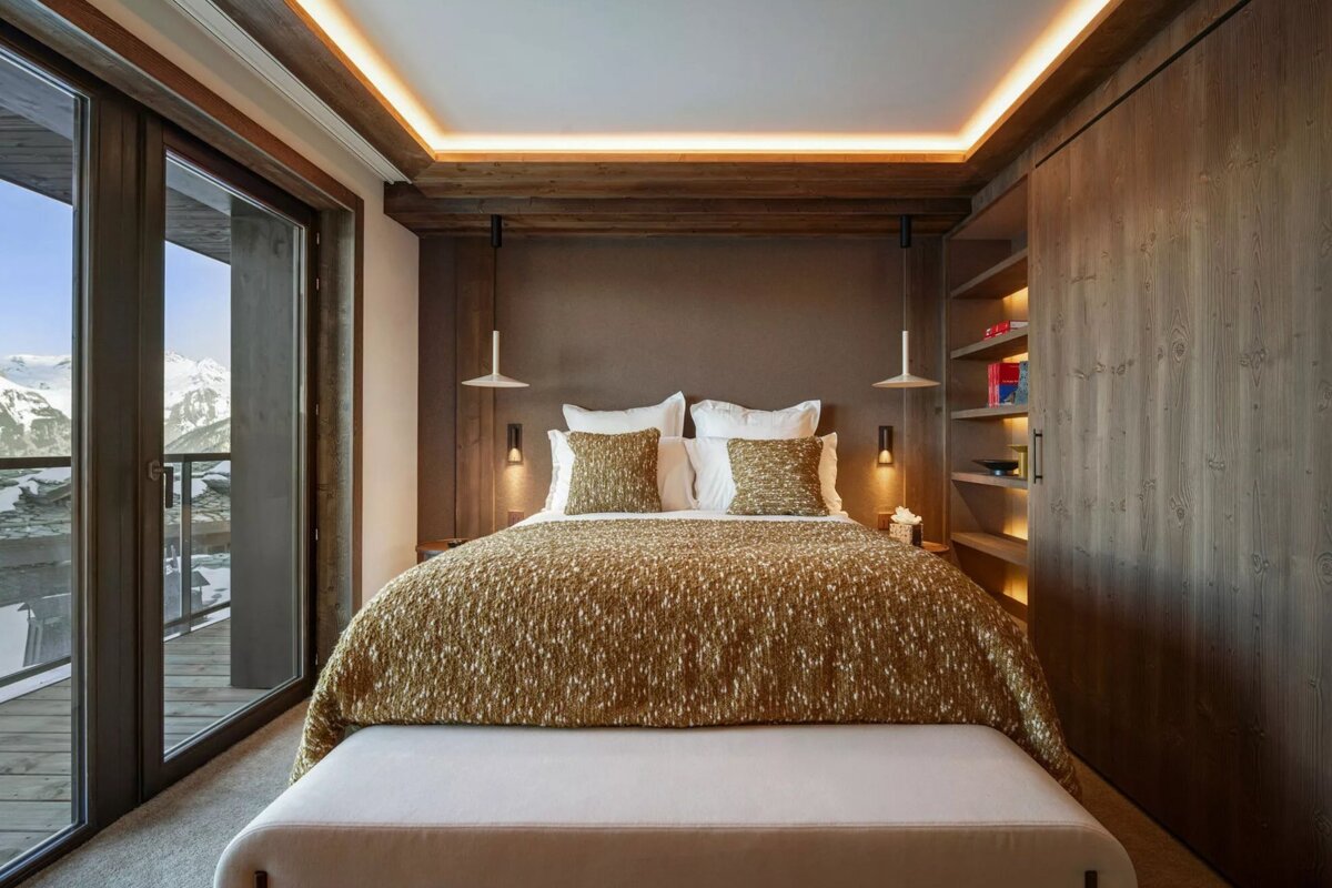 A modern bedroom with a golden bedspread, warm lighting, wood accents, and floor-to-ceiling windows offering a snowy mountain view.