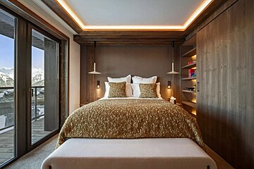 A modern bedroom with a golden bedspread, warm lighting, wood accents, and floor-to-ceiling windows offering a snowy mountain view.