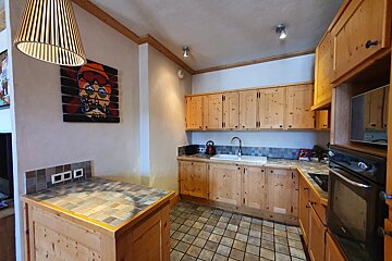 A rustic wooden kitchen with tiled floors and counters, featuring light wood cabinets, a modern hanging lamp, and a vibrant skull artwork.