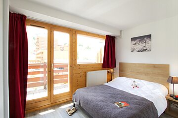 A bright bedroom features a bed with a wooden headboard, large wooden-framed windows leading to a balcony, and red curtains. Ski art adorns the wall.