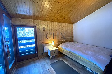 A cozy bedroom with a wooden bed, sloped wood-plank ceiling, and a glass door opening to a winter view.