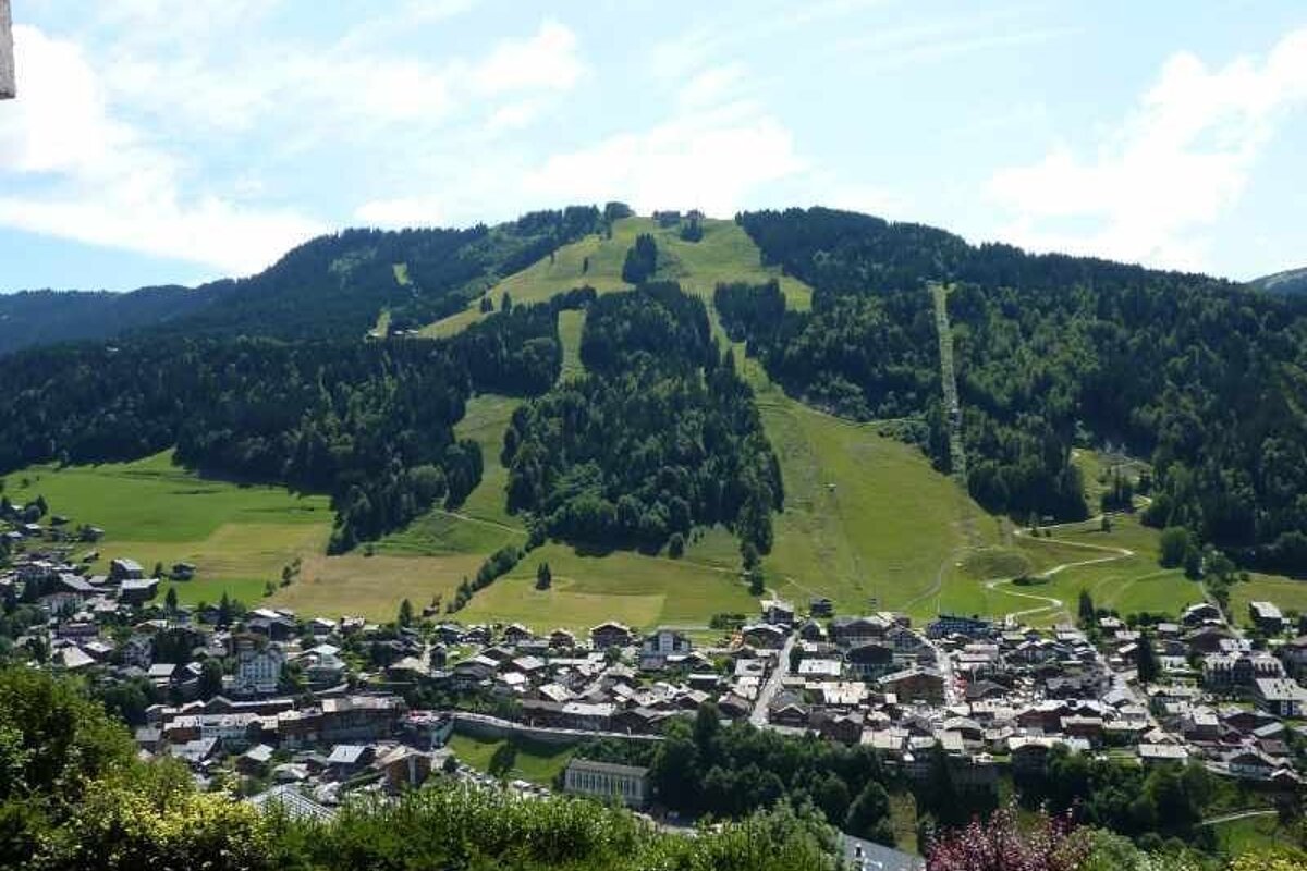 An alpine village nestled at the base of a lush green mountain with visible summer ski slopes.