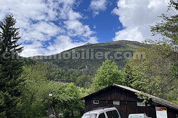 A lush green mountain under a blue, cloudy sky, with a dark wooden building and part of a silver car in the foreground.