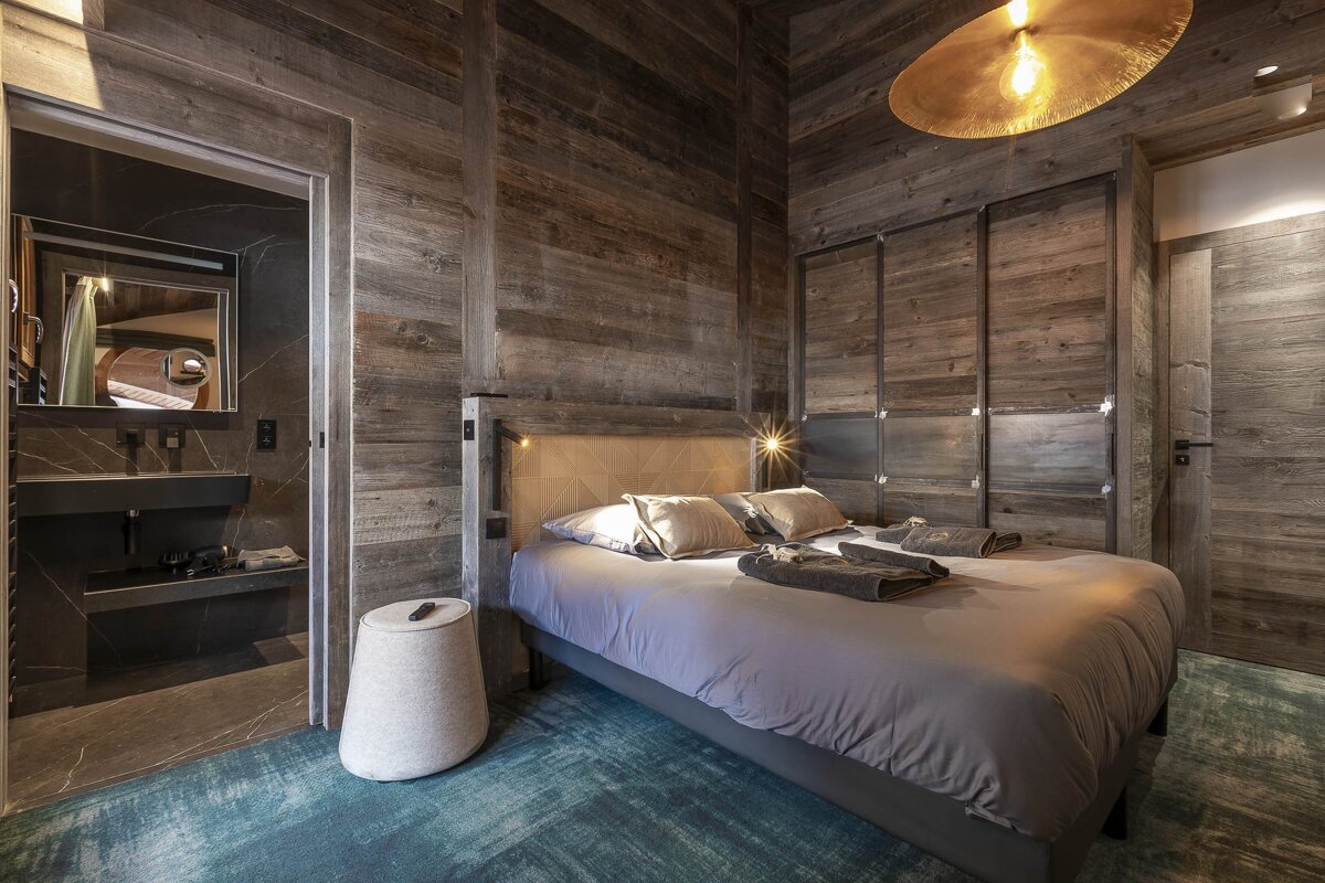 A modern bedroom featuring rustic wood walls, a gray bed with bright reading lights, a blue patterned carpet, and an adjacent bathroom with a black vanity.