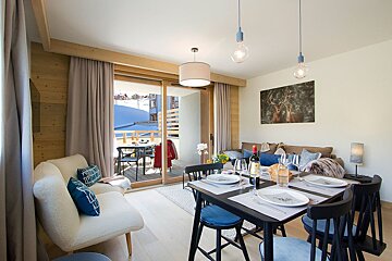 A cozy, open-plan living-dining room with a set table and sofa, featuring wood accents and a balcony overlooking a snowy landscape. Modern alpine style.