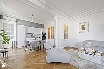 Chic, modern open-plan living room and kitchen with herringbone floors, white walls, minimalist grey furniture, and elegant ceiling moldings.