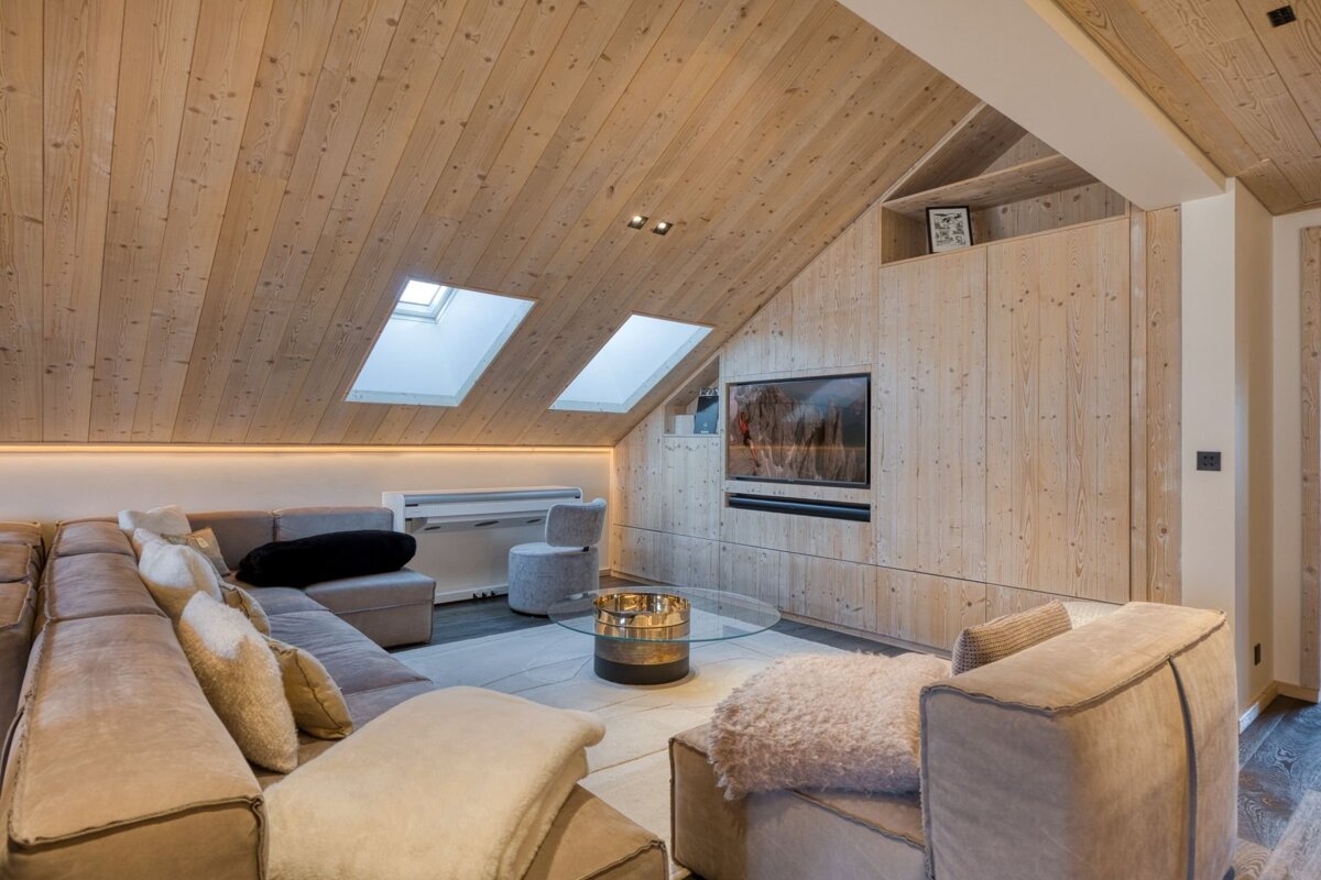 Cozy, modern attic living room with light wood paneling on walls and slanted ceiling, featuring skylights, a large L-shaped sofa, and a wall-mounted TV.