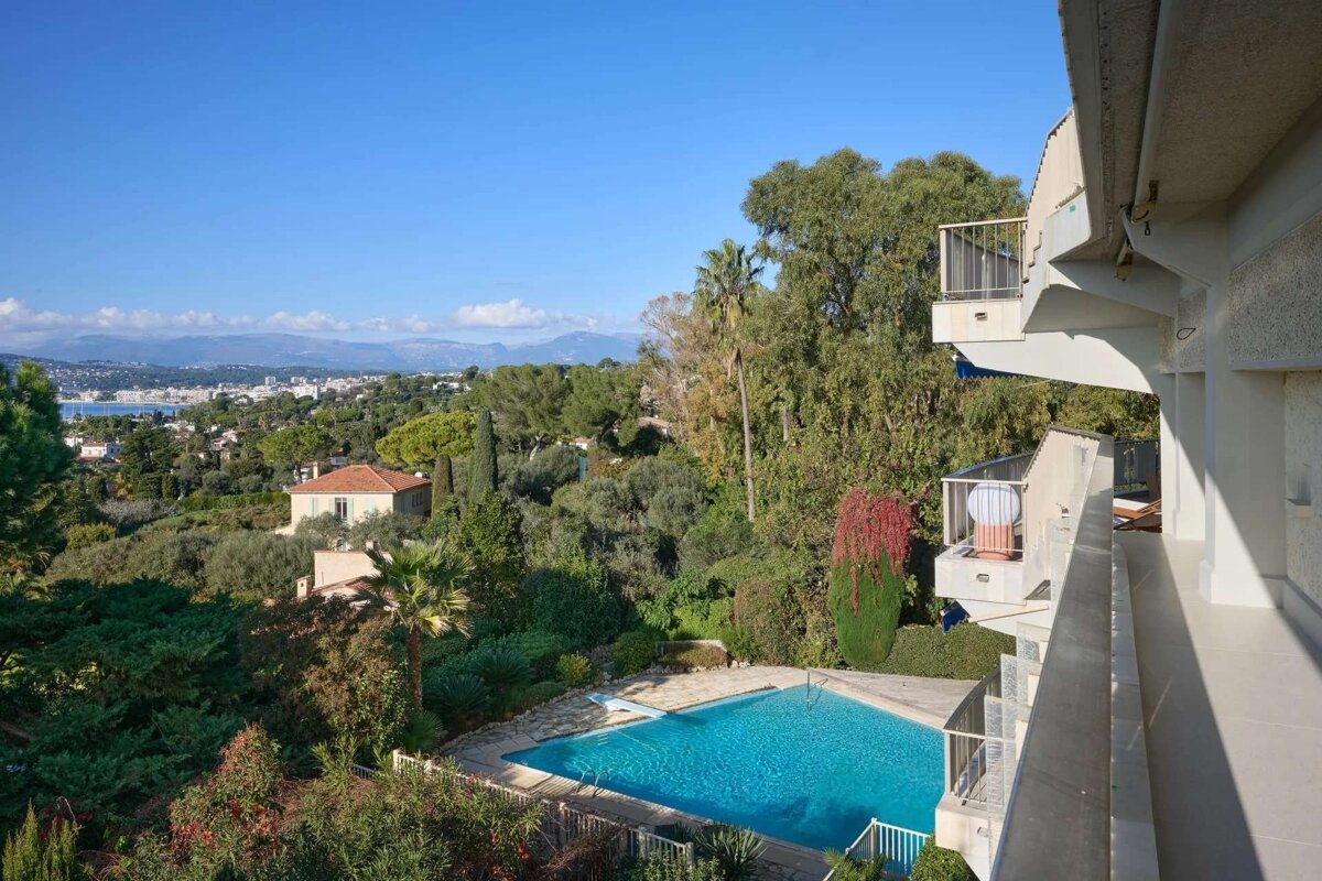 Balcony view of a sparkling pool nestled in lush greenery, with a distant coastal town and mountains under a clear blue sky.