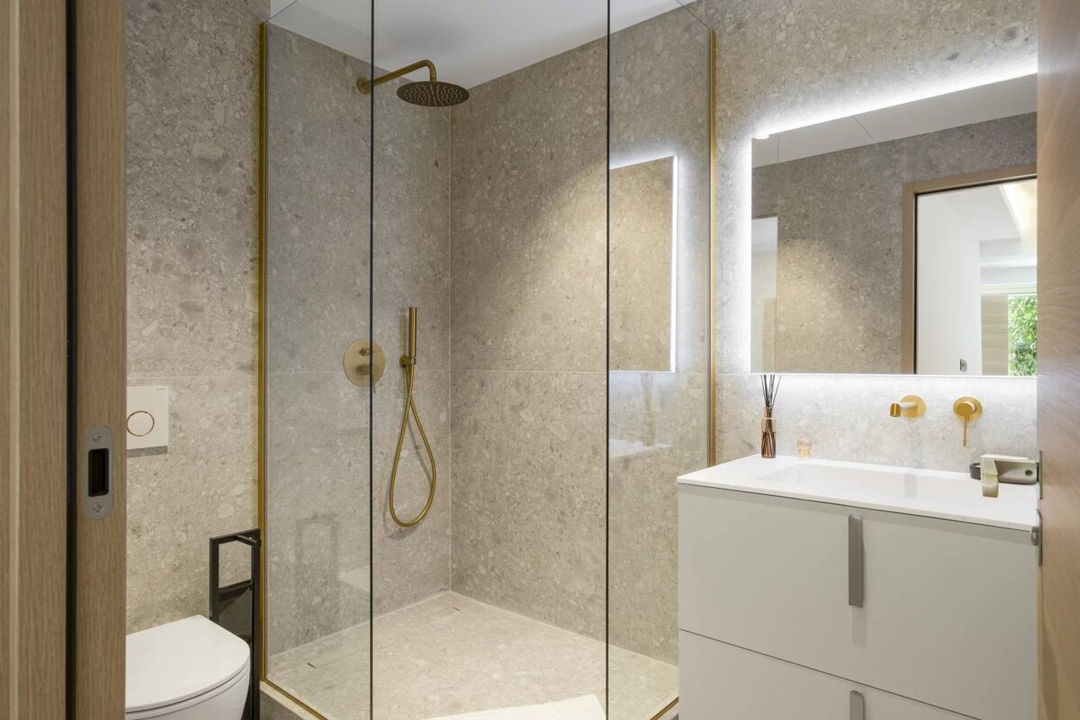 Modern bathroom with terrazzo tiling, a glass walk-in shower with gold fixtures, a white vanity, and a backlit mirror.