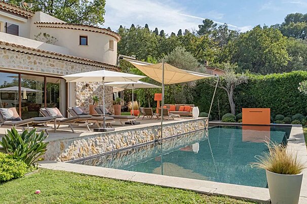 A charming stone villa with a dark-tiled pool, sun loungers, and an outdoor lounge under a canopy, all set in a lush, sunny garden.