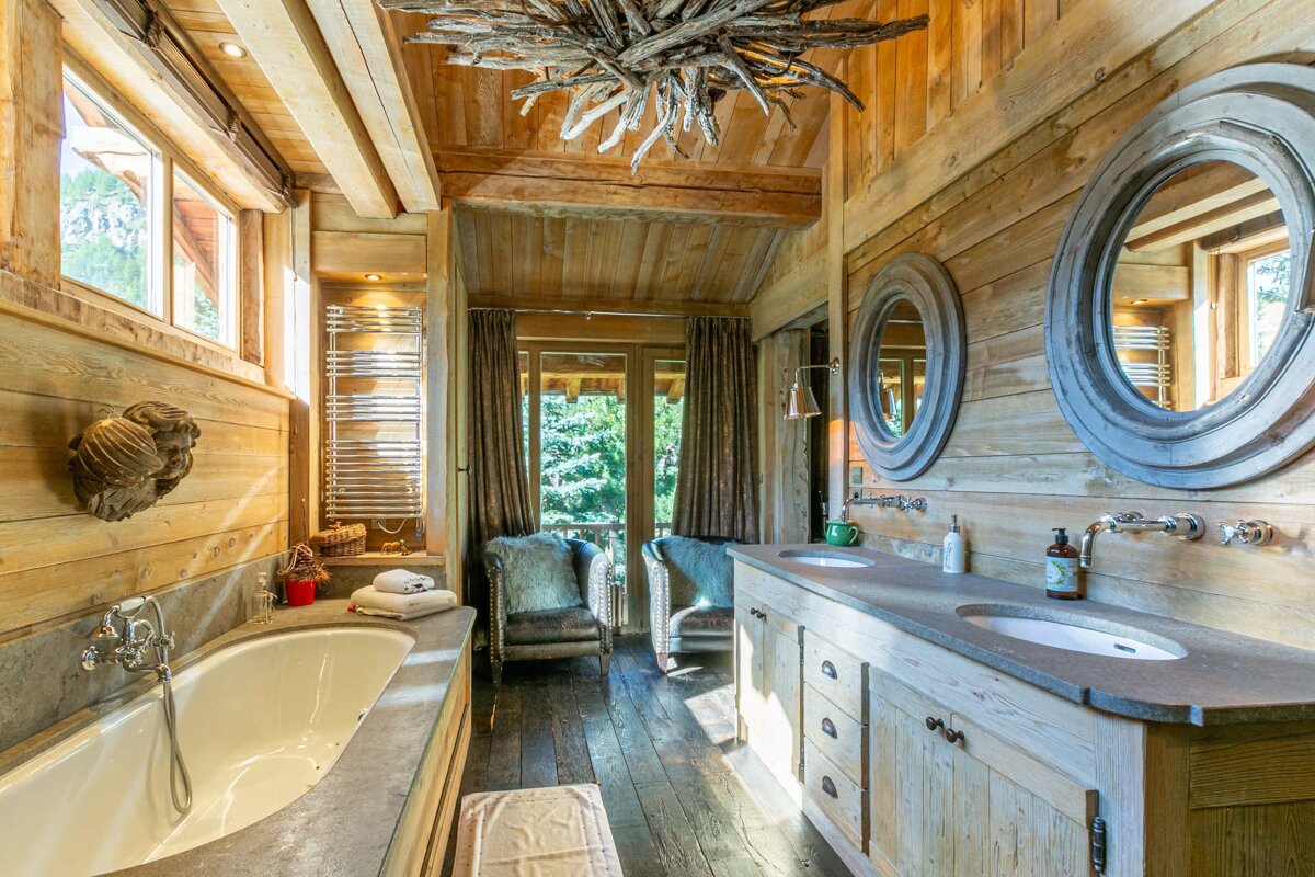 A luxurious, rustic wooden bathroom with a tub, double vanity, two furry chairs, a driftwood chandelier, and a forest view.