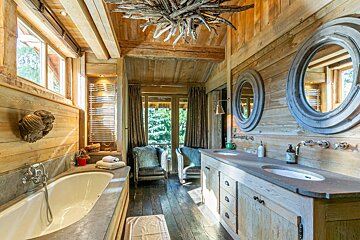 A luxurious, rustic wooden bathroom with a tub, double vanity, two furry chairs, a driftwood chandelier, and a forest view.