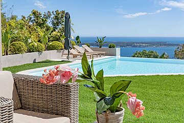 A luxurious infinity pool overlooking a beautiful ocean bay, framed by green lawn and tropical plants under a clear sky. Wicker furniture and a pink flower accent the foreground.