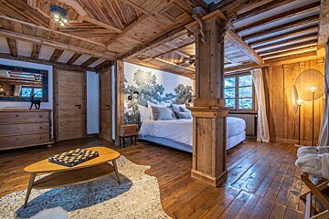Rustic wooden bedroom with a large bed, tree-patterned accent wall, exposed beams, and a chessboard on a coffee table.