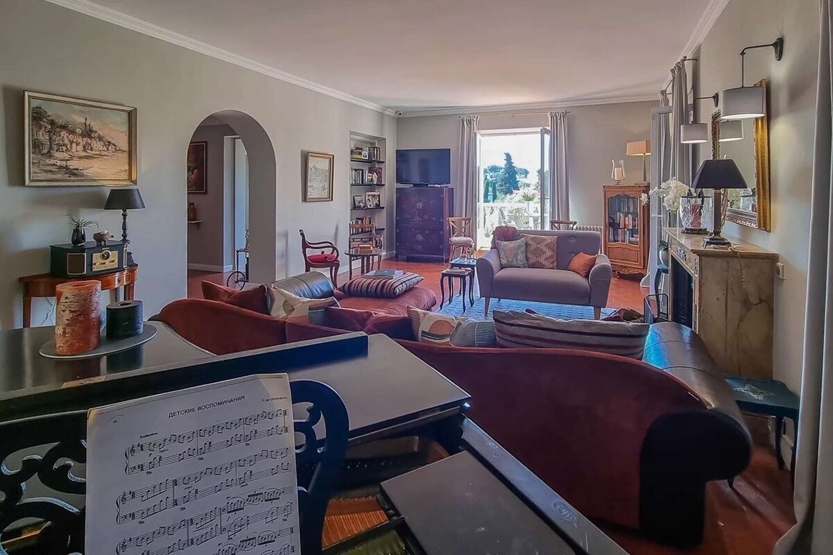 A spacious, elegantly furnished living room with a grand piano, various seating areas, and a balcony overlooking greenery.