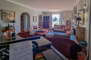 A spacious, elegantly furnished living room with a grand piano, various seating areas, and a balcony overlooking greenery.