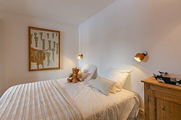 A bright, cozy bedroom features a single bed with white linens, a teddy bear, cow-themed wall art, and two warm wall lights.