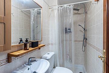 A compact bathroom with tiled walls, a sink with a mirror and wooden shelf, a toilet, and a shower enclosed by a clear polka-dot curtain.