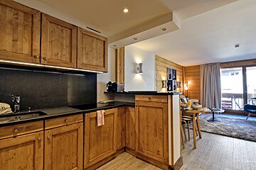 Cozy open-plan kitchen and living space with natural wood cabinets, dark counters, and wood-paneled walls, leading to a bright dining area and balcony.
