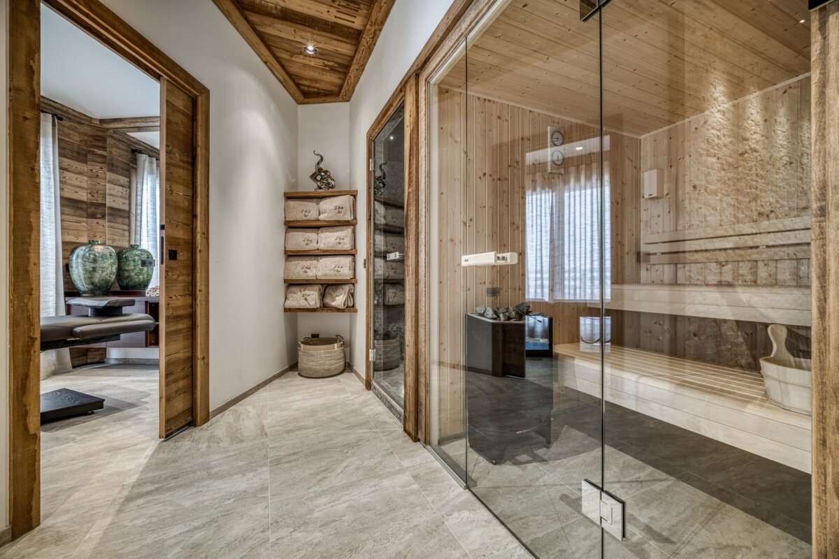 A modern spa area features a glass-front sauna, a tiled hallway with wooden accents, and a glimpse into a massage room with a table.