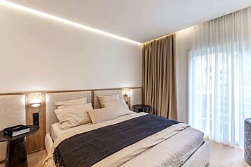 A modern, light-colored bedroom featuring a bed with integrated lighting, a dark throw, and full-length sheer and opaque curtains by a window.