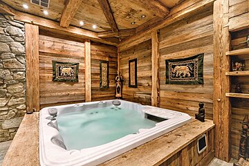 A cozy, rustic spa room with a bubbling white hot tub, surrounded by warm wooden walls, exposed beams, and a stone accent wall. Elephant tapestries and statues adorn the space.