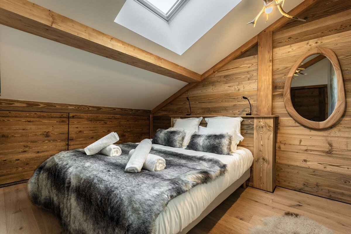 Warm, wood-paneled attic bedroom with a skylight, exposed beams, and a cozy bed adorned with a plush gray fur throw. Rustic charm.