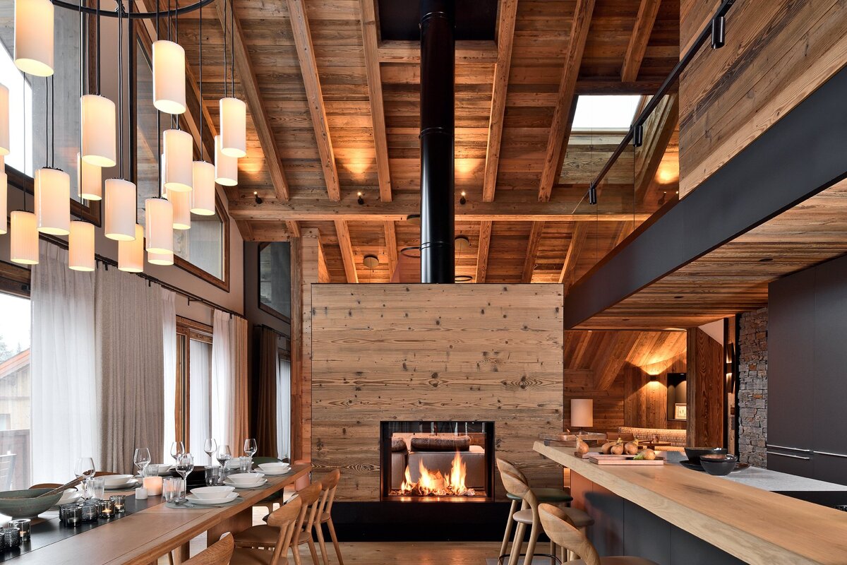 Modern chalet interior with a central wood-clad fireplace, a long dining table, elegant chandelier, and high wooden beamed ceilings.