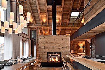 Modern chalet interior with a central wood-clad fireplace, a long dining table, elegant chandelier, and high wooden beamed ceilings.
