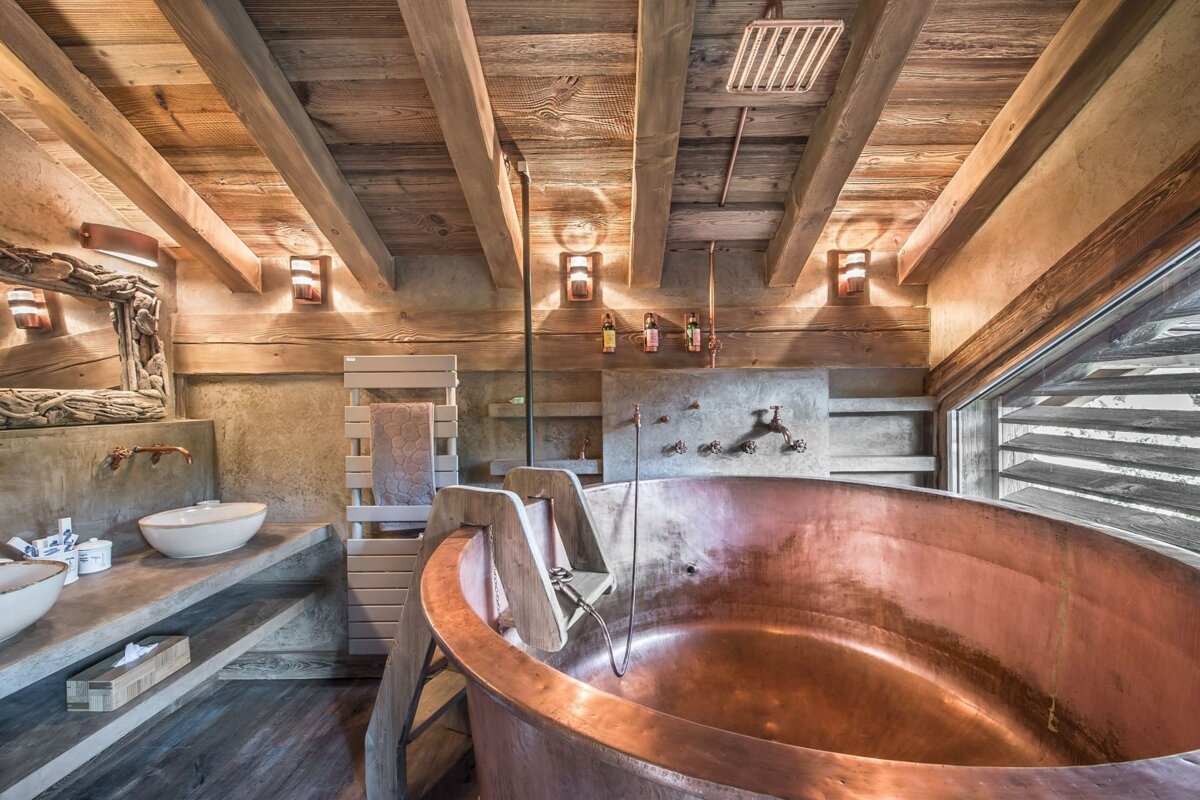 A luxurious rustic bathroom features a large copper bathtub, exposed wooden beams, concrete walls, and modern fixtures under warm lighting.