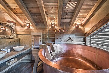 Cozy, rustic bathroom with a large copper tub, exposed wood beams, and stone walls. Industrial fixtures complete the warm, cabin-like feel.
