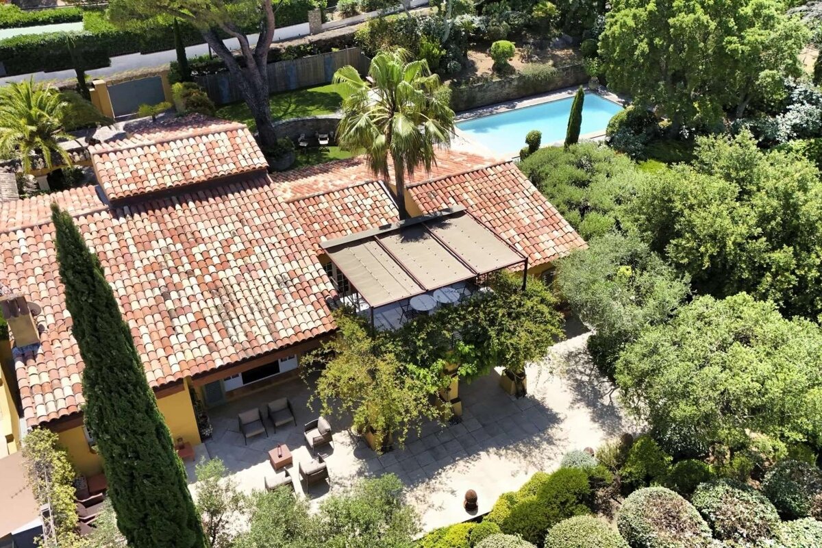 Aerial view of a house with a red tiled roof, surrounded by a lush garden, trees, and a blue swimming pool.