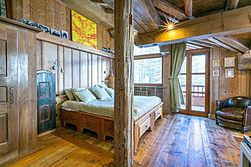 Rustic alpine chalet bedroom featuring warm wooden walls, exposed ceiling beams, a cozy bed, a leather armchair, and a glass balcony door.