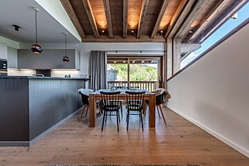 Modern dining area featuring a wooden table with black chairs, exposed timber ceiling beams, a kitchen counter, and sliding glass doors.