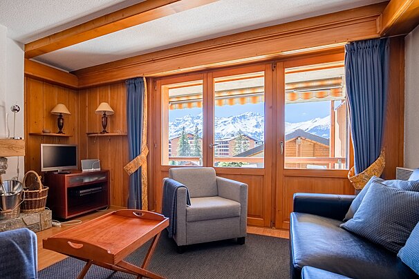 Cozy chalet living room with wood paneling, an armchair, and large glass doors opening to a scenic view of snowy mountains.