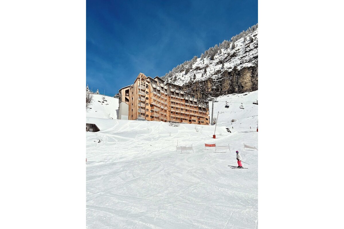 A skier descends a sunny mountain slope with a large wooden building, snow-covered mountains, and a clear blue sky in the background.
