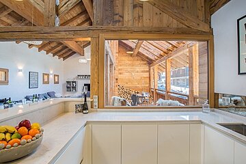 Modern white kitchen with a bowl of fruit, looking through a large interior window into a rustic wooden chalet.