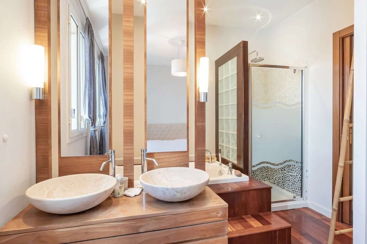Modern bathroom with twin stone vessel sinks on a wooden vanity, mirrors reflecting a bedroom, a glass shower, and wooden flooring.