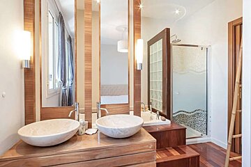 Modern bathroom with twin stone vessel sinks on a wooden vanity, mirrors reflecting a bedroom, a glass shower, and wooden flooring.