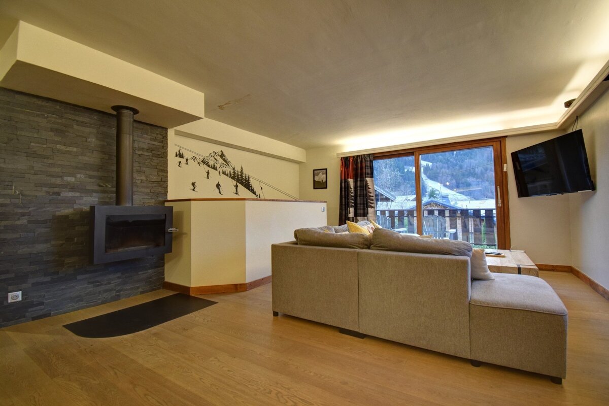 A modern living room featuring a gray sectional sofa, wood floor, wall-mounted fireplace, and a large window with a mountain view. A skiing mural decorates the wall.