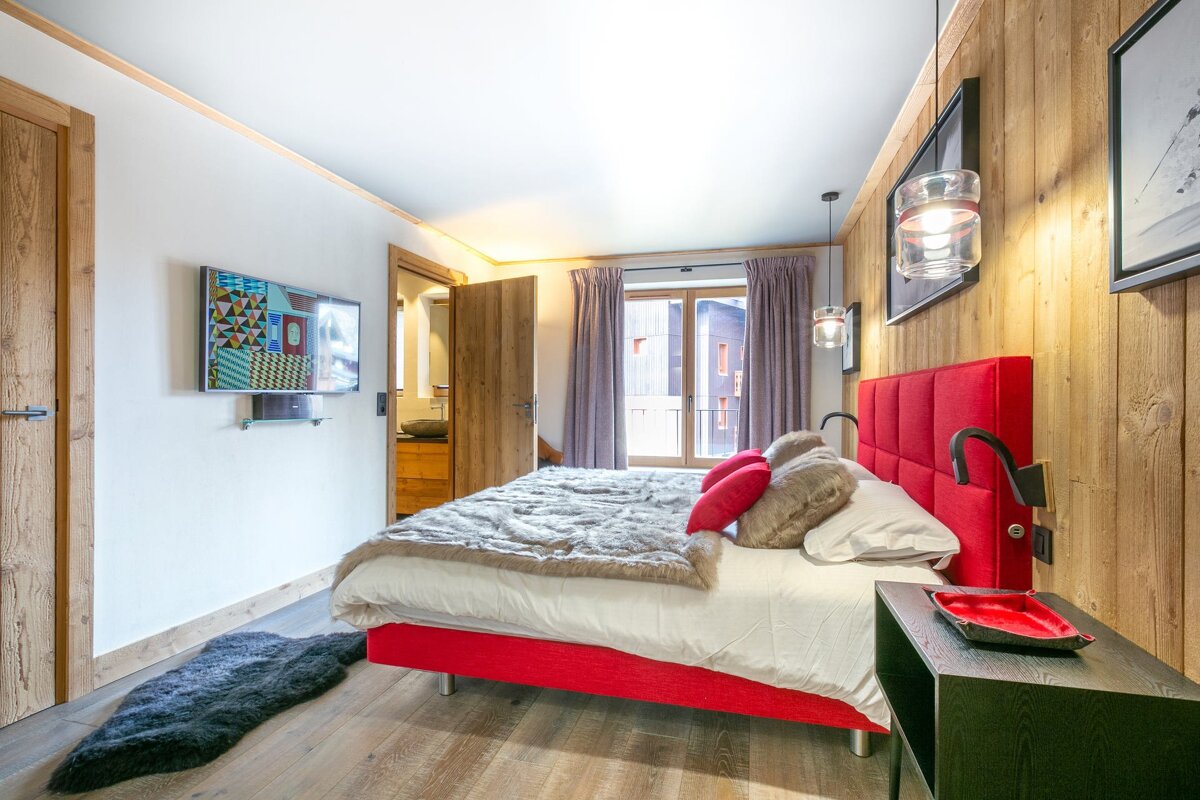 Modern, cozy bedroom with a vibrant red bed, wood-paneled wall, mounted TV, fur throw, and a balcony view.